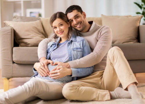 pregnancy, love and care concept - happy man hugging pregnant woman and making hand heart gesture at home